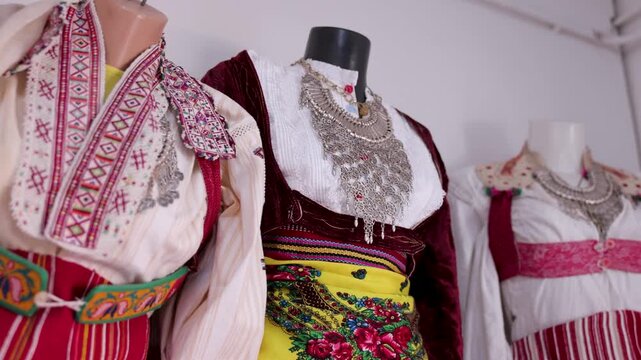 Slow pan across three traditional Albanian folk costumes displayed on mannequins. Colorful embroidery, woven fabrics, and ornate jewelry highlight cultural heritage.
