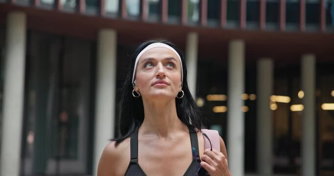 A young beautiful female athlete in sportswear and a backpack goes for a workout in an urban park among high-rise buildings