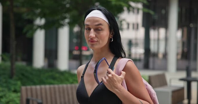 A young beautiful female athlete in sportswear and a backpack goes for a workout in an urban park among high-rise buildings