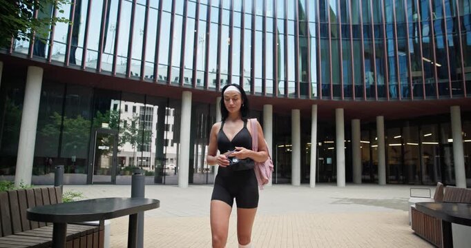 A young beautiful female athlete in sportswear and a backpack goes for a workout in an urban park among high-rise buildings