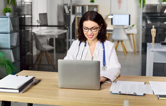 Doctor with laptop working online in clinic. Female physician in office uses computer and stethoscope to manage telehealth and update patient files. Concept shows modern digital healthcare.