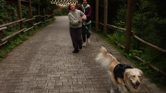 Mother and daughter have fun running with golden retriever on a forest path in the evening