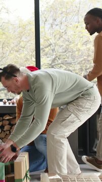 Vertical video: Bending to pick green gift, man wearing reindeer headband arranging gifts at home