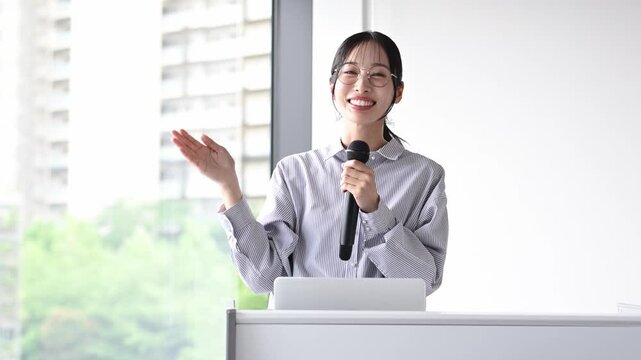 会議室でマイクを持ってプレゼンテーションを行う30代の日本人ビジネスパーソンの女性
