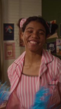 Vertical portrait of African American female teenager jumping and waving pom-poms at camera while dancing in bedroom. Youth lifestyle marketing, mental health campaigns, or social media content