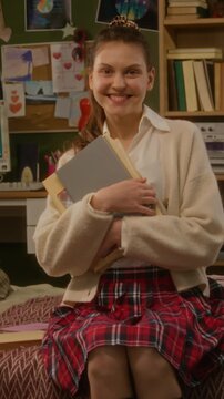Vertical portrait of Caucasian female teenage high school student holding books, smiling at camera in student room or bedroom. Footage is perfect for back-to-school promos and e-learning ads, vertical