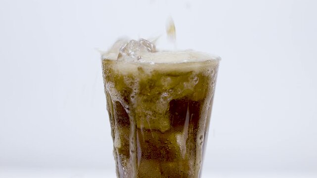 A dramatic close-up of cola exploding and splashing high out of a glass with ice cubes against a white background.