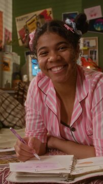 Portrait of African American teenager in pink pajamas lying on bed, writing in diary and smiling at camera. Clip is ideal for youth lifestyle, mental health, or journaling apps, vertical