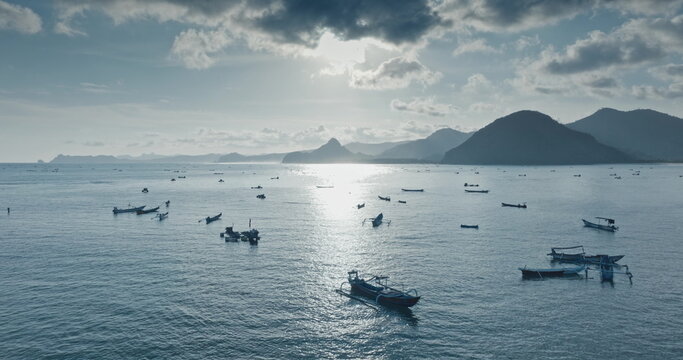 Indonesian fishing boats float on calm ocean water at Selong Belanak Beach, Lombok, reflecting warm sunlight under cloudy sky with tropical mountains rising in background. Blue evening toning filter