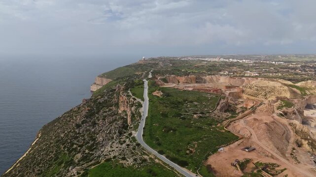 At Malta's Dingli Cliffs, the rocky coastline and surrounding rural fields with a winding road and nearby quarry site extend toward the Mediterranean Sea under an overcast sky in a drone view.