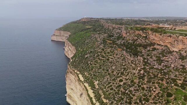 In Malta's Dingli Cliffs area, an aerial view shows the sheer limestone escarpment meeting the Mediterranean Sea along a rugged, vegetation-lined coastline under overcast winter skies.