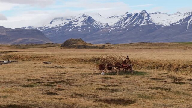 Antique wheel rake abandoned in field surrounded by snowy mountains, aerial arc