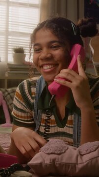 Vertical video of African American teenager lying on bed, talking on retro pink corded telephone, laughing and twirling cord. Clip is ideal for nostalgia marketing or youth lifestyle campaigns