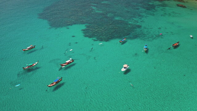 Traditional longtail boats and yacht floating on the clear turquoise tropical sea over visible coral reef, with people swimming and enjoying the vacation in Phuket, Thailand. Aerial view drone flight