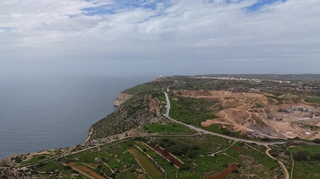 In Dingli, Malta, the Dingli Cliffs rise above a rocky Mediterranean coastline bordered by terraced fields, vegetation, and a rural road, seen in a wide perspective from a drone.