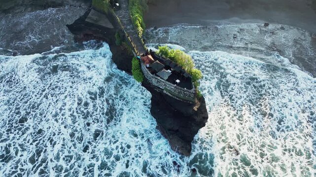 White surf and powerful ocean waves surrounding a sacred Hindu temple on a rocky coastal outcrop in warm morning light, overhead drone footage.
