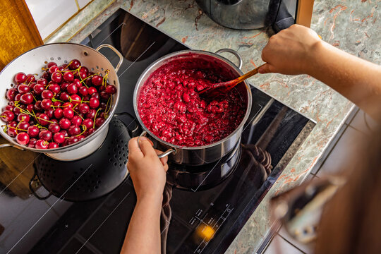 Hands stirring a pot of bubbling cherry jam on a modern induction stovetop