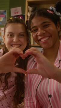 Portrait of Caucasian teenager and African American girl wearing pajamas, smiling and making heart shape with hands during sleepover in bedroom. Youth lifestyle and diversity campaigns, vertical