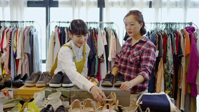 Two cheerful young asian women small business owners and young female customers choosing secondhand shoes and clothing items in a modern boutique, preparing stylish merchandise for online retail sale
