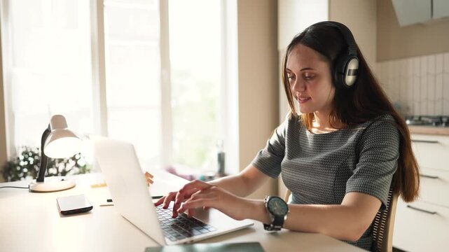 Young woman typing on modern laptop keyboard. Female worker wears large headphone. Student doing remote work at home. Freelancer uses computer in sunny office. Smiling woman finishes laptop work.