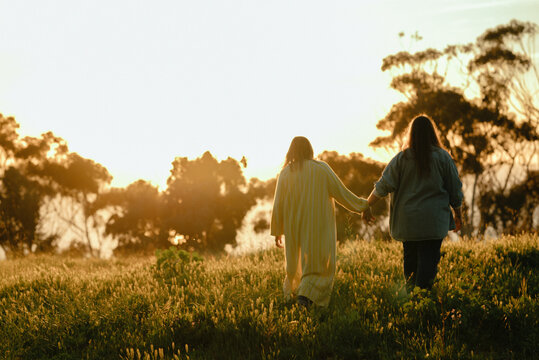 Two women holding hands walking through a field at sunset