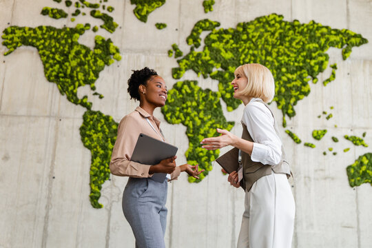 Two businesswomen discussing sustainable global strategy in modern office with green world map wall