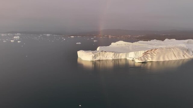 Majestic iceberg floats past a Greenland town with a rainbow