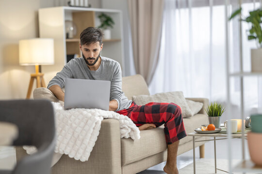 Caucasian man working on laptop at home on couch in casual pajama pants