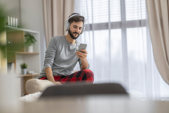 Caucasian man with headphones using smartphone at home in casual loungewear relaxing on bed