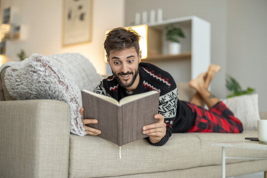 Caucasian man reading book on couch at home, relaxed and surprised expression in cozy living room