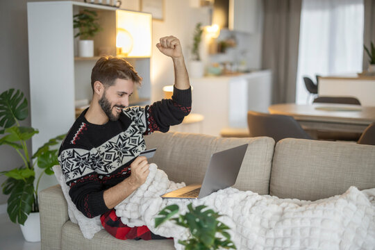 Man celebrating online purchase on laptop at home holding credit card (Caucasian)