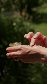 Closeup of adult hands poised over open cosmetic jar in sunlit light, with blurred green foliage backdrop