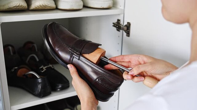 Person inserts a wooden shoe tree into a leather loafer beside a wardrobe. Crease prevention and shape retention improve long term footwear storage.