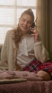 Vertical of Caucasian teenage girl in high school uniform sitting on bed, talking on retro cordless phone and smiling, vertical. Clip is perfect for nostalgic lifestyle or retro technology ads