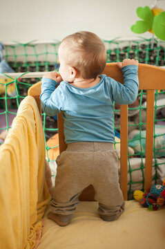 Back view of tiny explorer embraces the morning light, standing tall and steady, grasping the polished wood of the crib, eager for play