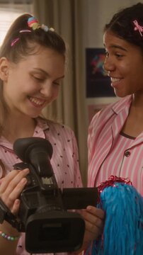 Vertical video of Caucasian and African American teenage girlfriends in pink pajamas reviewing footage on video camera screen. Clip is ideal for youth vlogging and social media marketing campaigns