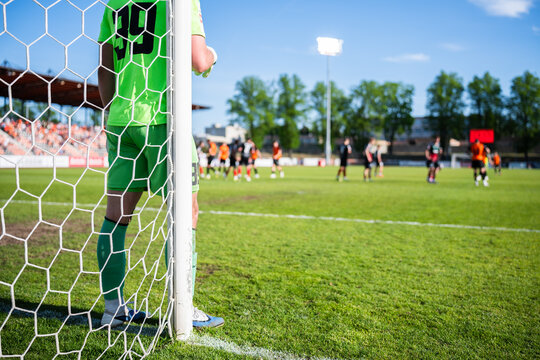 Rear view of a soccer goalkeeper in a green kit standing by the goalpost and watching the game through the net.