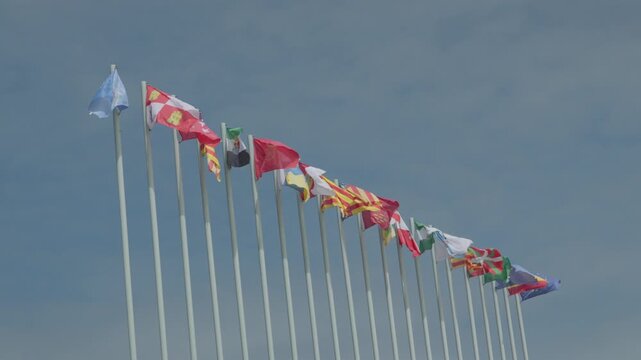 Low-angle video clip of a long row of colorful flags fluttering in the wind on tall flagpoles against a clear blue sky. Captured at the Philippe Cousteau Anchor Museum in Salinas, Asturias, Spain.