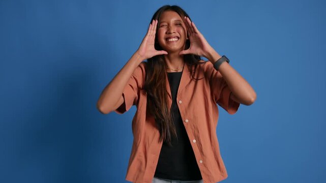 A young woman stands in front of a blue background, shouting towards the camera, announcing or calling someone with both hands at the sides of her mouth.