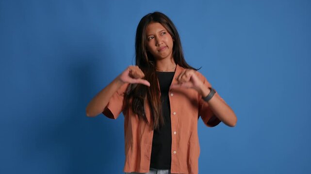 A young woman stands in front of a blue background, looking disappointed or rejecting something, unhappy, while showing a thumbs down.