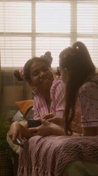 Vertical video of African American teenager and Caucasian teenager in pajamas lying on bed, sharing cookies and watching television. Ideal for streaming service ads or snack brand marketing