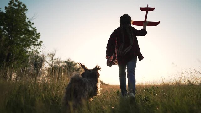 Girl running with toy airplane through meadow at sunset. Child holds toy plane high while running in grass field. Kid plays with airplane in countryside. Girl dreams of flying at golden hour.