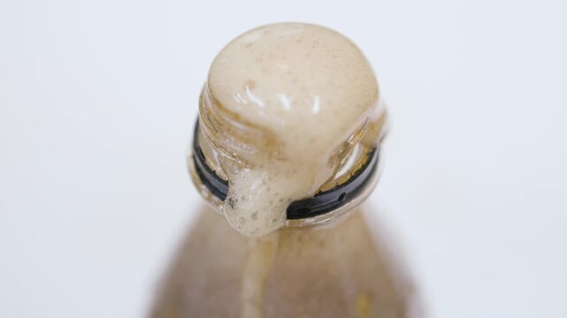 A close-up of foam pouring and overflowing from the top of a cola bottle against a white background.