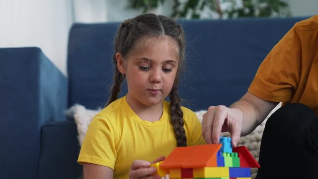 Mother and girl play together. Girl builds colorful toy house. Mother helps girl place plastic block. Family enjoys fun time inside living room. They construct bright block house. Toy brings joy.