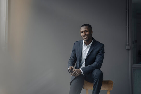 Portrait of smiling young businessman with smartphone sitting on backrest against grey background