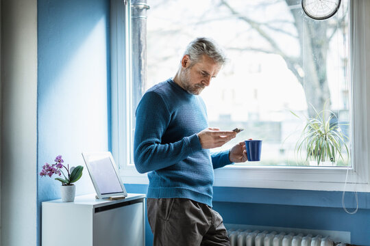 Mature man standing with coffee mug standing in living room looking at smartphone