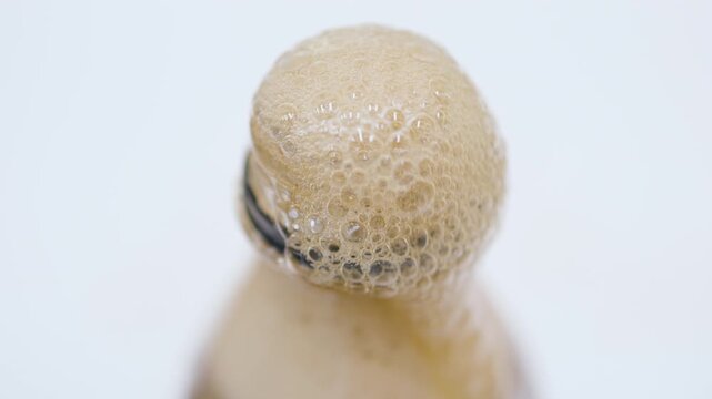A close-up of cola foam bubbling and overflowing dramatically from a bottle opening against a white background.