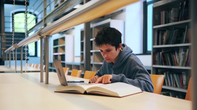 A focused teenage boy sits at a desk in a university library, reading a book and typing on a laptop. He is preparing for an exam or doing homework.