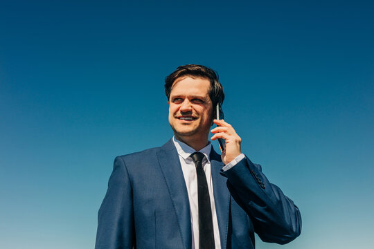Smiling businessman talking on smartphone outdoors against blue sky