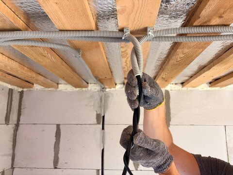 an electrician's hands installing electrical wiring in a corrugated conduit on a wooden ceiling during construction. Process of residential or commercial electrical rough-in and renovation.